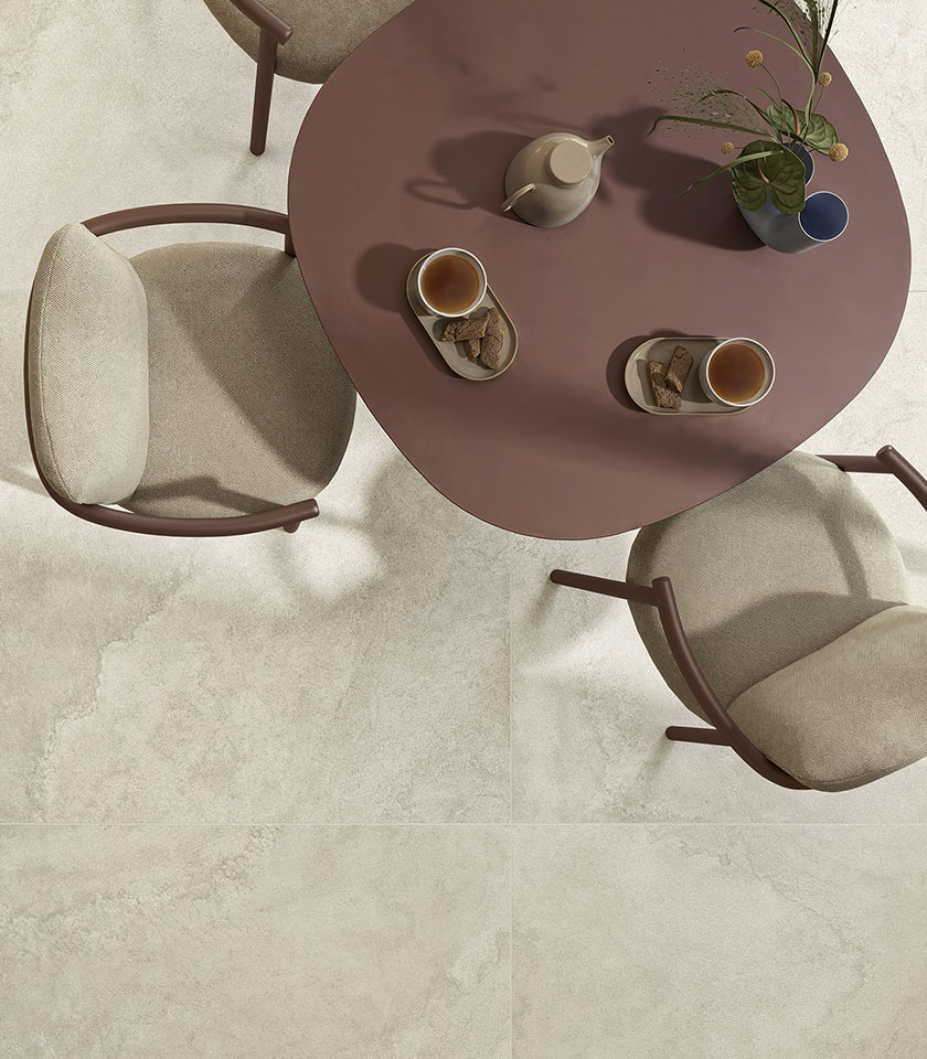 Terracotta-colored round table with tea service and beige upholstered chairs on a floor from the Pulse collection, with a stone effect in the Shell shade.